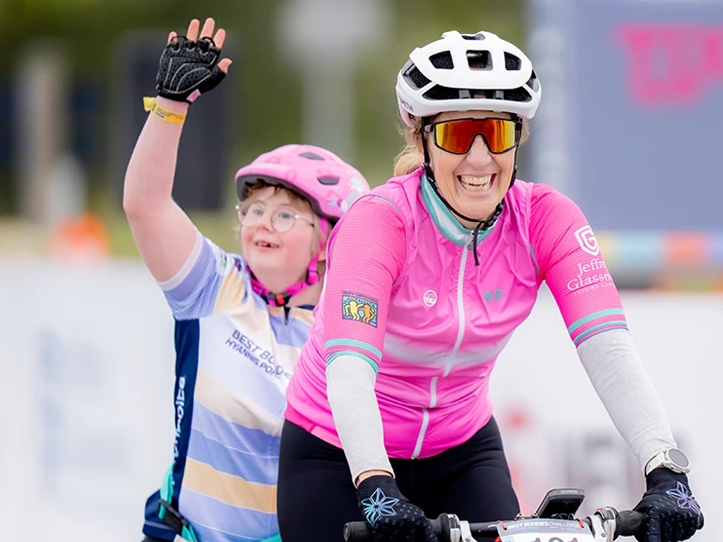 Two cyclists wearing helmets, one smiling and waving behind the other in a pink jersey