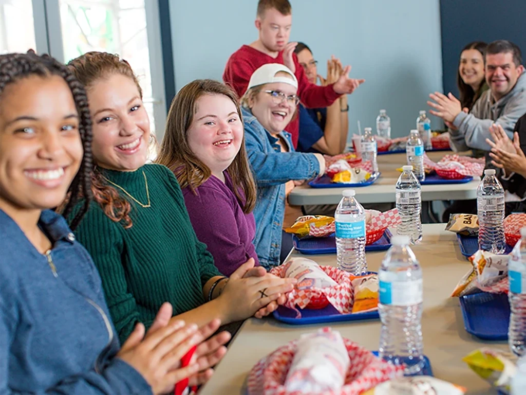 A group of people smiling and clapping around a table with meal trays and water bottles