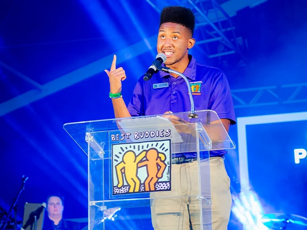 A young man speaking at a podium with a Best Buddies logo on the front